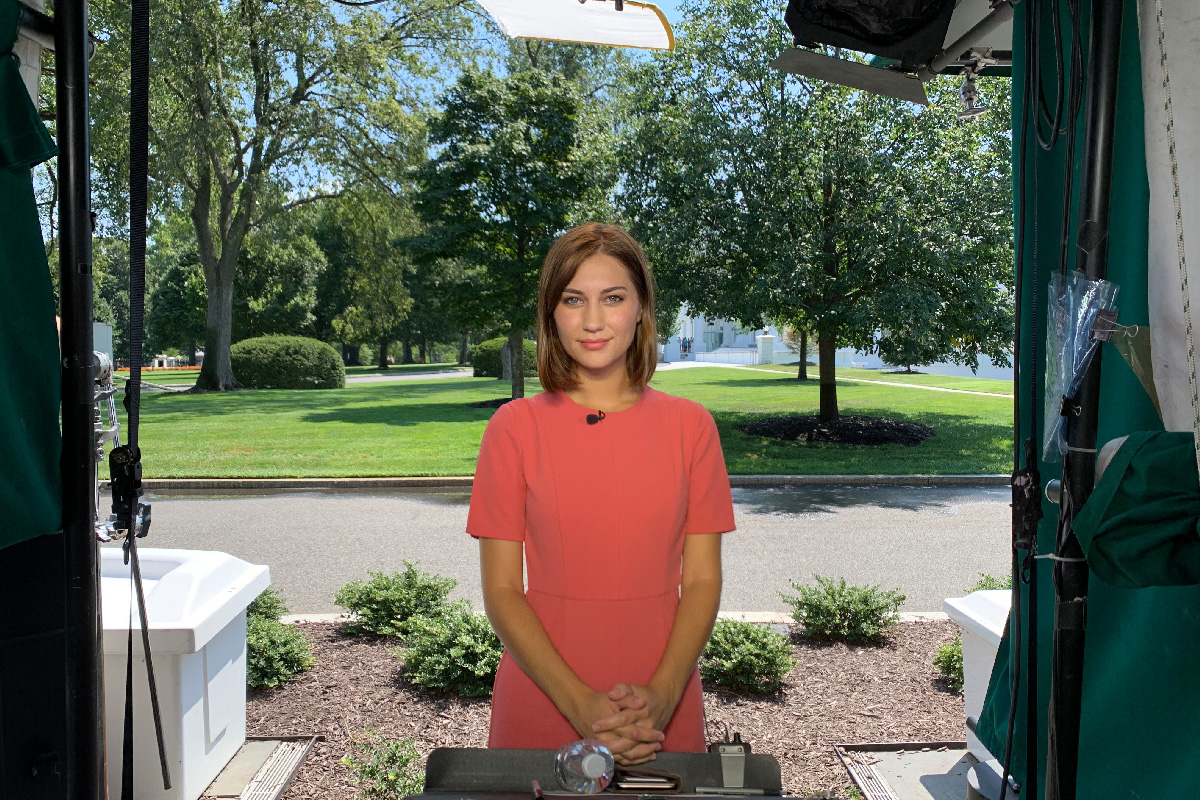 Woman stands before cameras preparing for a newscast in a pink dress.